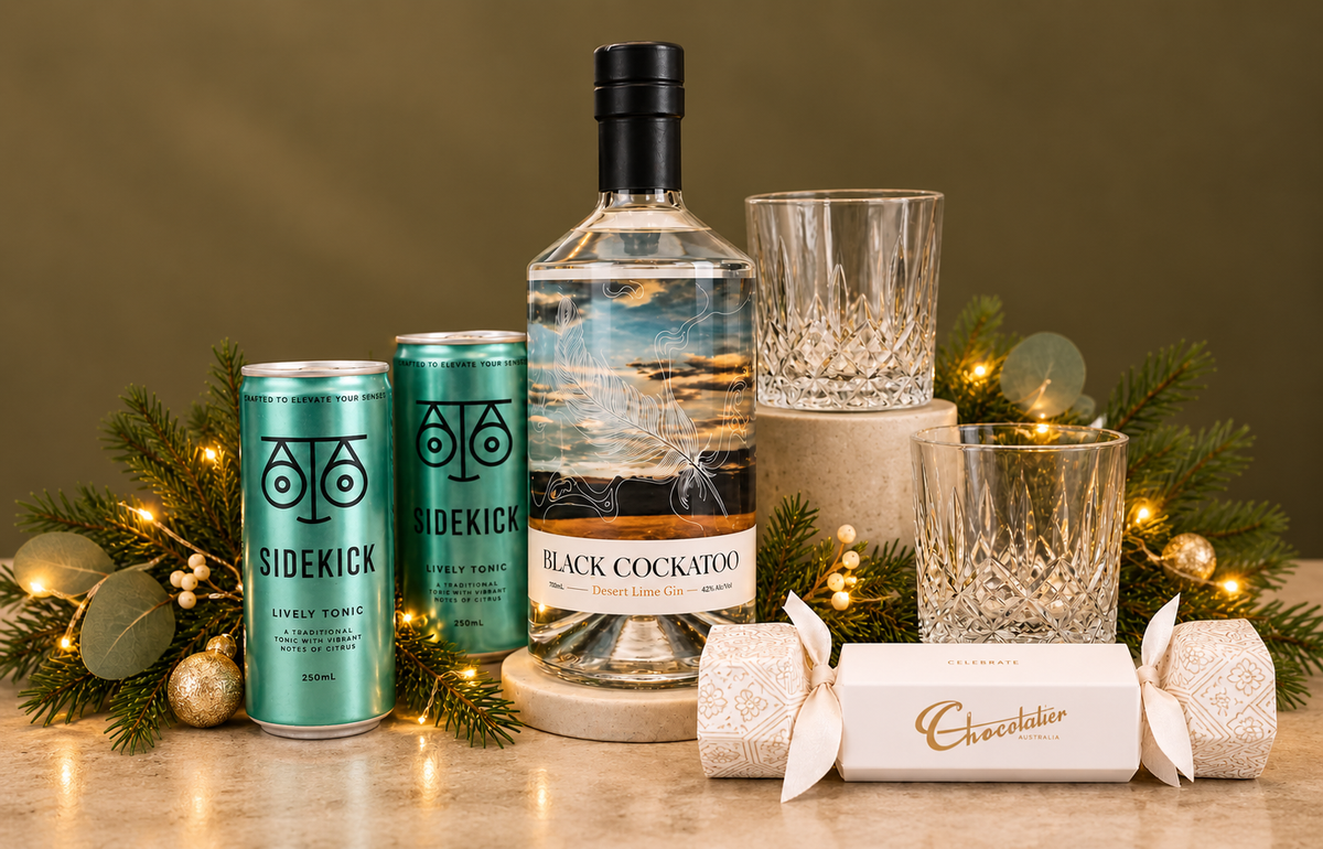 Bottle of gin, two glass tumblers, two sidekick mixers and a chocolate bon bon on green background with greenery and lights.