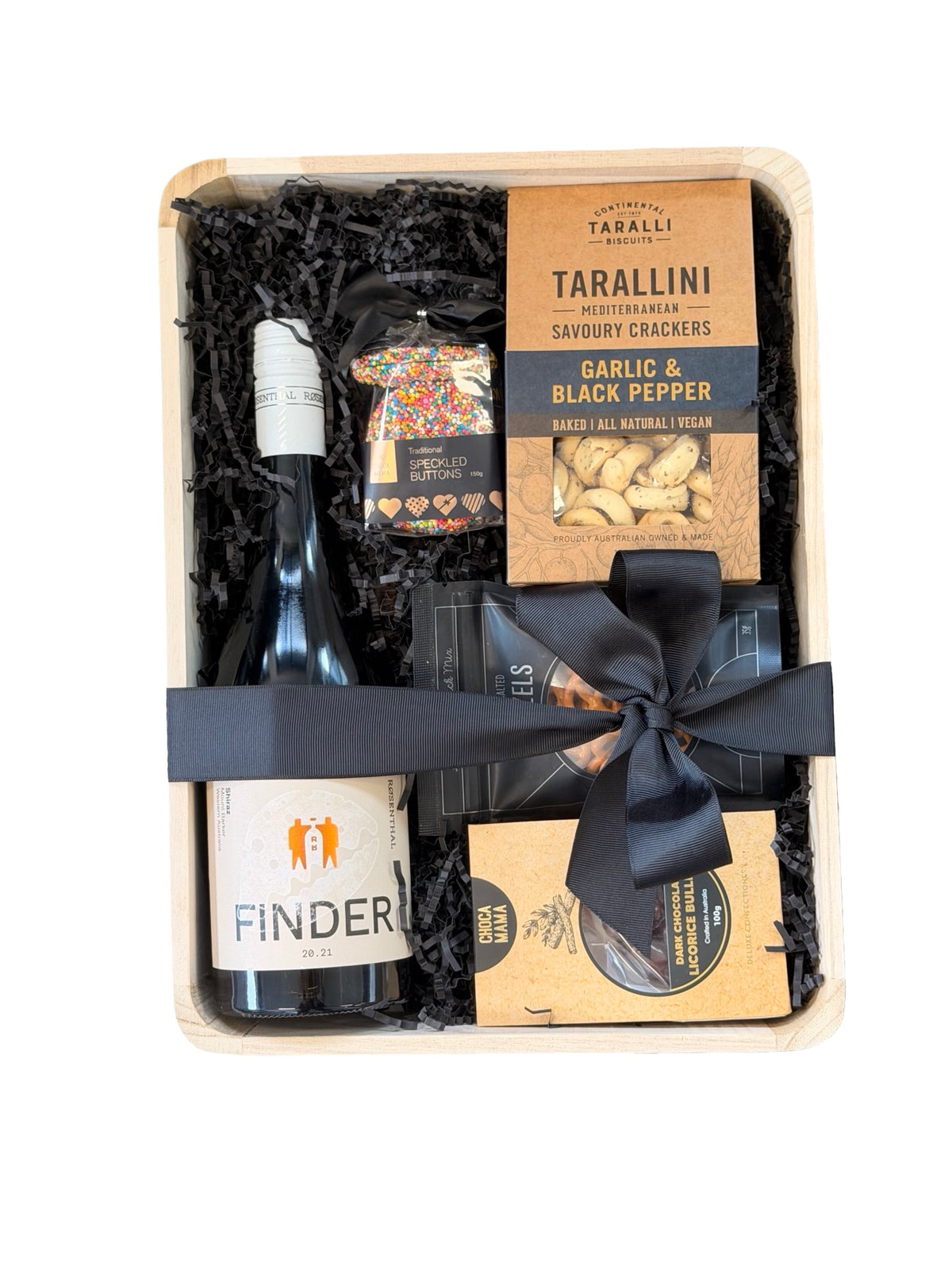 A curated gourmet gift box set in a light wood tray, filled with black crinkle paper shred. The set includes a bottle of Finder wine, Tarallini Savory Crackers, a bag of garlic and black pepper, and a small packaged item tied with a black ribbon.