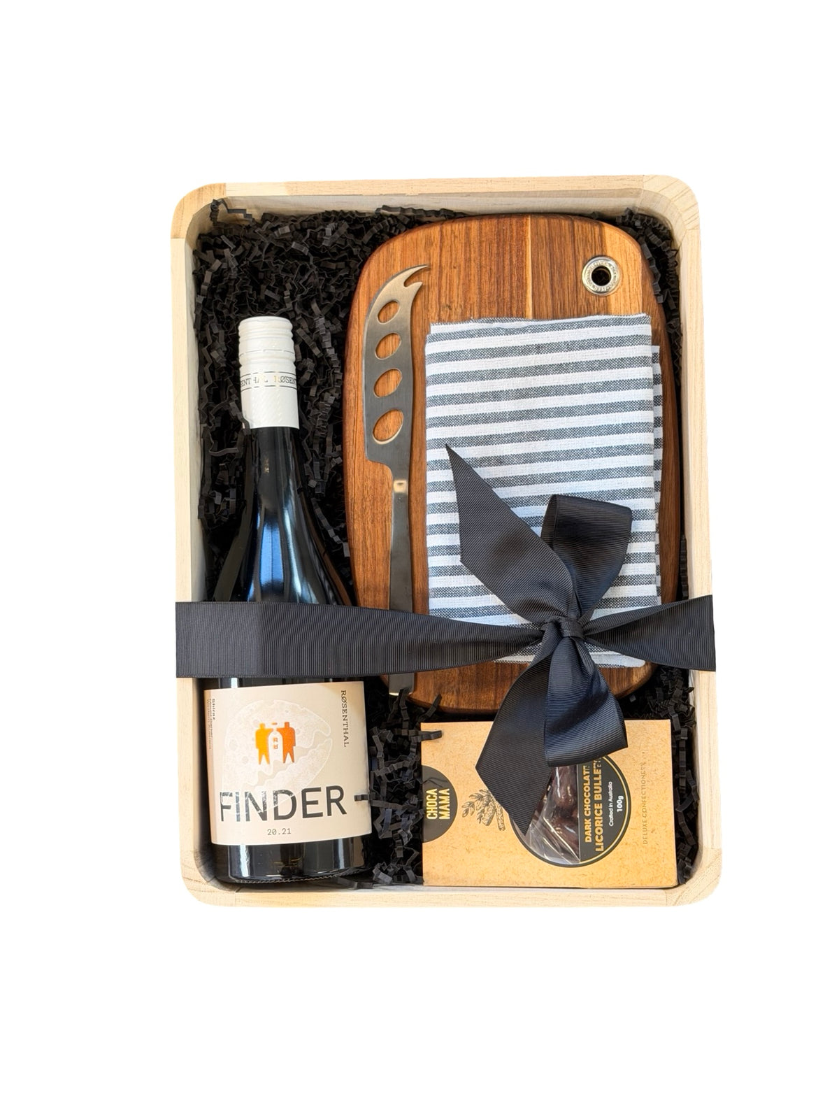 A curated gift basket set in a light wood tray with black shredded filler, featuring a bottle of Finder wine, a wooden cutting board set with a striped cloth and cheese knife, and a packaged food item tied with a black ribbon.