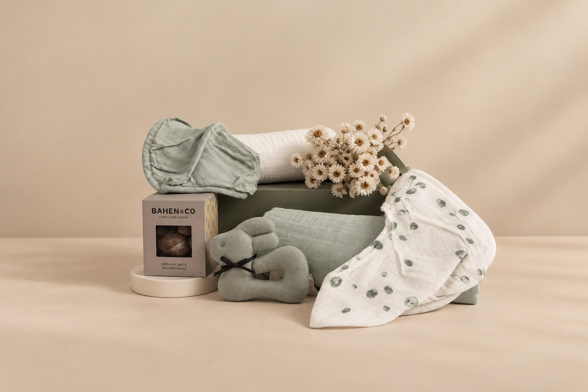 A collection of baby essentials including swaddles, bandana bibs, and a linen rattle, presented in a white magnetic box with a floral decoration.