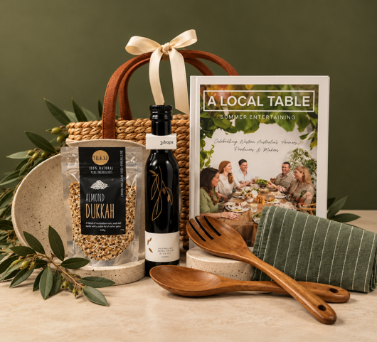 A styled gift basket arrangement on a light stone surface against a dark green wall. The set includes a woven basket with a cream ribbon, a bag of almond dukkah, a small dark bottle, a printed booklet titled 'A LOCAL TABLE Summer Entertaining', wooden serving utensils, and a folded green striped cloth, decorated with greenery and pinecones.