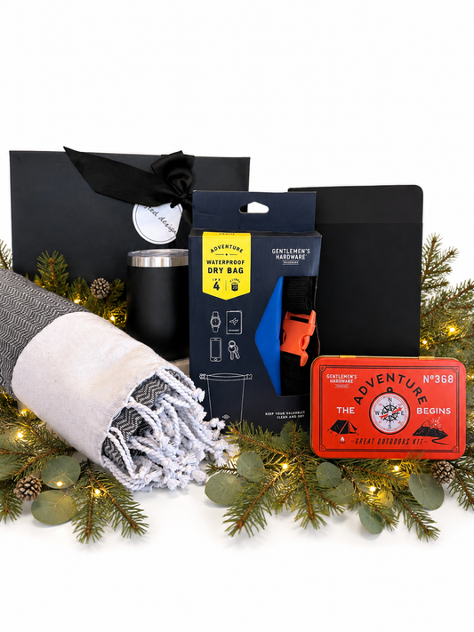 A curated gift set arranged on a white background with pine branches and fairy lights. The set includes a black gift bag with a black ribbon, a black travel dry bag, a black insulated tumbler, a blue and orange tent footprint, a red vintage-style ticket, and a folded gray-and-white fringed blanket.