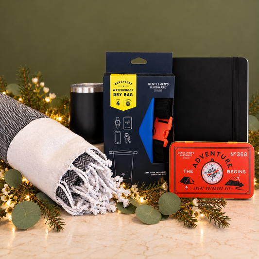A curated gift set arranged on a green background with pine branches and fairy lights. The set includes a black journal, a black travel dry bag, a black insulated tumbler, a blue and orange tent footprint, a red vintage-style ticket, and a folded gray-and-white fringed blanket.