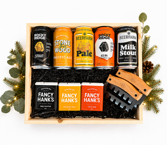 A curated gift box featuring five beverage cans from Rocky Ridge and Beerfarm, three Fancy Hanks seasoning packets, and two wooden-handled meat grills nestled in black shredded paper inside a light wood crate, decorated with eucalyptus and fairy lights.