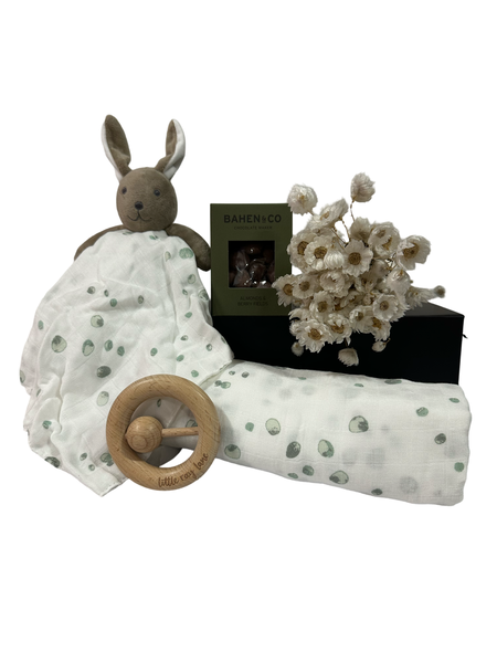 A baby gift set including a swaddle, comforter, wooden teether, and chocolates, presented in a white gift box with decorative flowers.