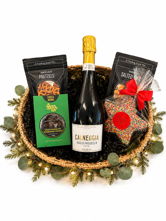 A holiday gift basket arrangement on a white background, featuring a bottle of Cal Neggia Aglio Prosecco, a bag Pretzels, a bag of Salted Pretzels, and a star-shaped bag of sprinkles tied with a red bow, all set in a woven basket with greenery and fairy lights.