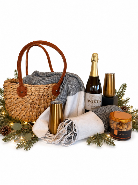 A curated gift basket arrangement on a white background featuring a woven straw tote with brown leather handles, a gray-and-white patterned throw blanket, a bottle of Poét champagne, two gold-toned cocktail glasses, a small jar of candied nuts, and pine greenery with fairy lights.