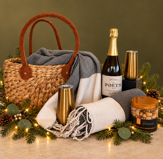 Harvest basket with two Turkish towels, bottle of sparkling, good insulated tumblers and jar of salted nuts. On green background with greenery and lights