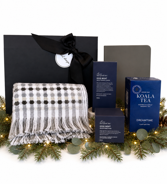 A curated wellness gift set arranged on a white background with decorative evergreen branches and fairy lights. The set includes a black gift box tied with a black ribbon, a gray-and-white fringed throw blanket, a dark blue box of Koala Tea Dreamtime tea, a dark blue box of Good Night body wash, and a dark gray box.