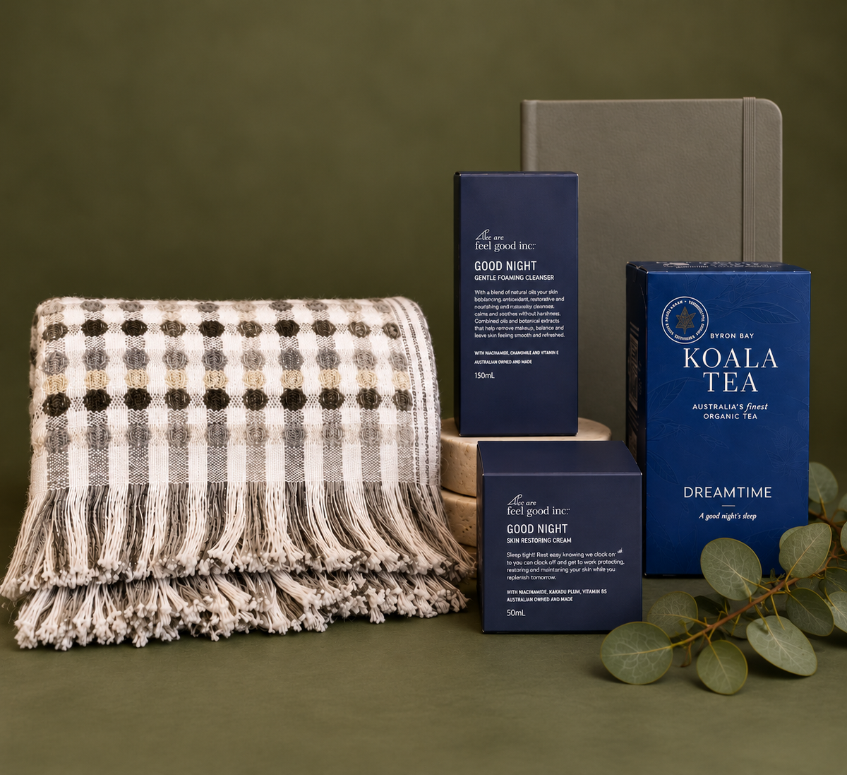 A curated wellness gift set arranged on a white background with decorative evergreen branches and fairy lights. The set includes a black gift box tied with a black ribbon, a gray-and-white fringed throw blanket, a dark blue box of Koala Tea Dreamtime tea, a dark blue box of Good Night body wash, and a dark gray box.