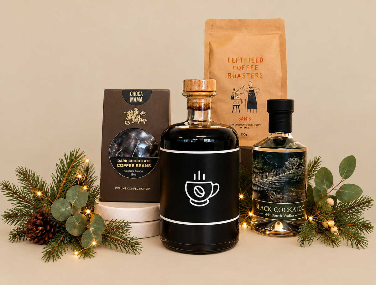 A curated gift set featuring a black gift bag with a black ribbon, a brown coffee roast pouch, a black chocolate coffee beans box, a dark liquid bottle with a coffee cup icon, and a black cocktail bottle, styled with greenery and fairy lights on a white background.