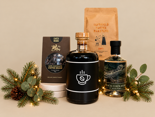 A curated gift set featuring a black gift bag with a black ribbon, a brown coffee roast pouch, a black chocolate coffee beans box, a dark liquid bottle with a coffee cup icon, and a black cocktail bottle, styled with greenery and fairy lights on a white background.