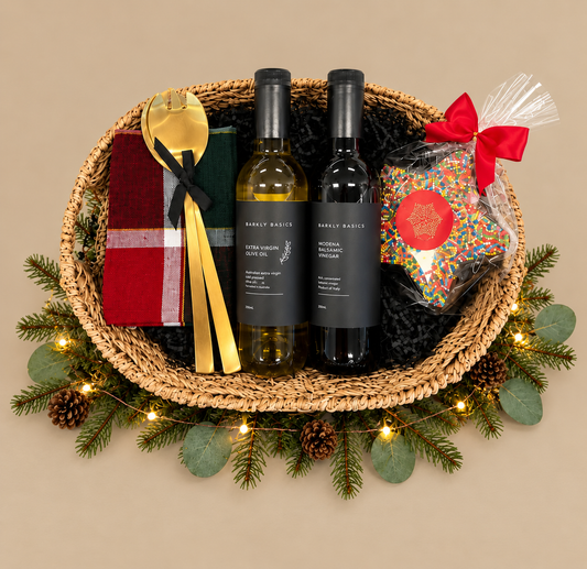 A holiday gift basket set from Simply Brisket, featuring two bottles of wine (one white, one red), a red and green plaid napkin, gold-colored cutlery, and a bag of sprinkles tied with a red bow, all arranged in a woven basket with a pine garland and fairy lights.