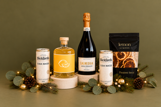 A curated gift set arranged on white pedestals in front of a black gift box with a black ribbon. The set includes a bottle of Calneggia Asolo Prosecco, a bottle of citrus soda water, two cans of Bickfords Soda Water, and a black bag of lemon limone sparkling soda water, all decorated with green pine garlands, eucalyptus, and warm fairy lights on a white background.
