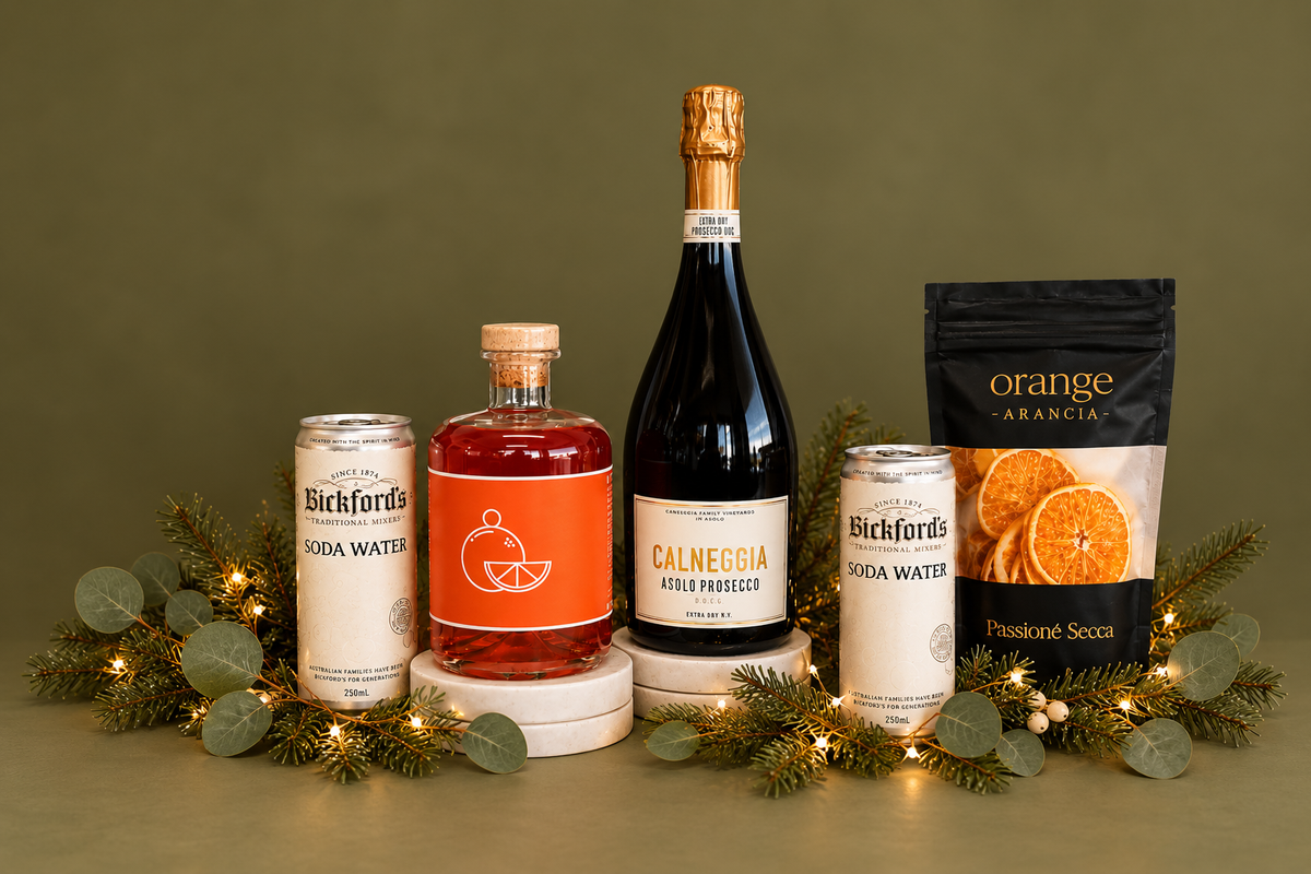 A curated gift set featuring a bottle of Calneggia Amaro Prosecco, a bottle of orange-flavored soda water, two cans of Bickfords soda water, a black pouch of orange-flavored confection, and a black gift box, all styled with green eucalyptus, pine branches, and warm fairy lights on a white background.
