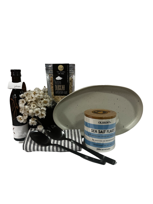 A gift box arranged with a ceramic jar of sea salt flakes, a serving platter, a black salad server, a dukkah spoon, a striped tea towel all against a white background.