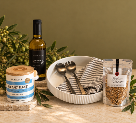 A gift box arranged with a ceramic jar of sea salt flakes, a serving platter, a black salad server, a dukkah spoon, a striped tea towel all against a white background.