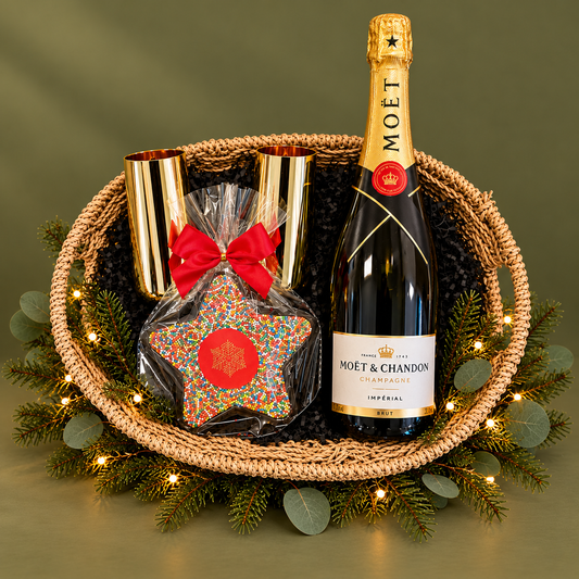A festive gift basket arrangement on a white background, featuring a bottle of Moët & Chandon champagne, a gold-capped bottle opener, a star-shaped chocolate covered in multicolor sprinkles with a red bow, and a woven basket decorated with greenery and fairy lights.