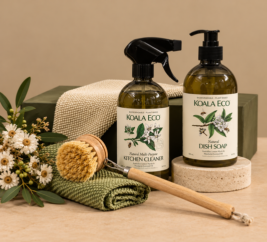 Koala Eco natural multi-purpose kitchen cleaner spray bottle and dish soap pump bottle displayed with a wooden-handled brush, folded green cloth, and olive branches on a beige surface.