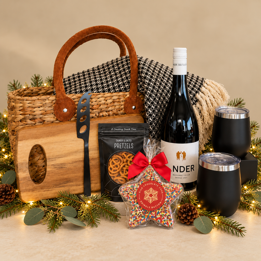A holiday-themed gift basket arrangement on a white background, featuring a woven straw basket with brown leather handles, a wooden cutting board with a black-handled knife, a bottle of wine, two black insulated tumblers, a black package of gourmet pretzels, a red bow, and a decorative star ornament, styled with pine branches, eucalyptus, and fairy lights.