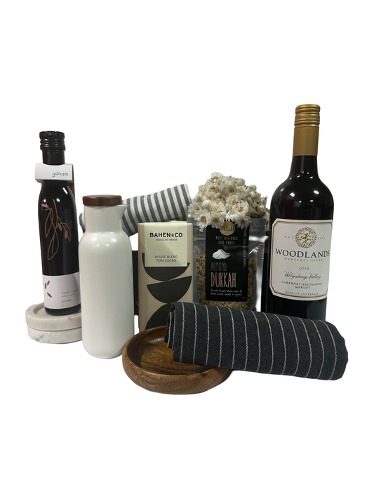 A gift box containing a ceramic jug, a small wooden bowl, black and white striped tea towels, a bottle of wine, olive oil, sukkah, and chocolate, all arranged on a matte black surface.