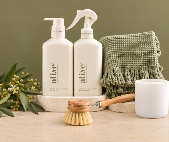 Two white bath and body products from the brand 'alive' on a marble tray, styled with a green fringed towel, a wooden scrubber, a white mug, and dried flowers against a green background.