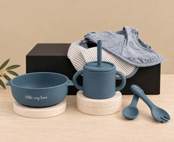 A coordinated baby feeding set in matte slate blue, including a bowl with a straw, a handled tumbler, a spoon, and a fork, styled with dried flowers, a striped cloth, and a black box on a warm beige surface.