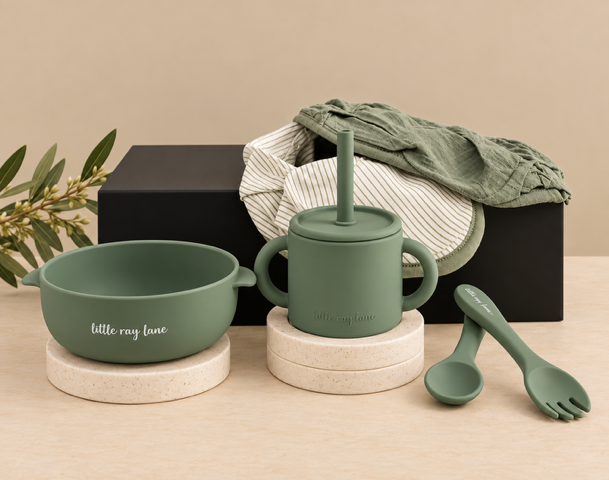 A coordinated baby feeding set in matte sage green, including a bowl, handled cup with lid and straw, and utensils, styled with dried flowers, a striped cloth, and a black box on a warm beige surface.