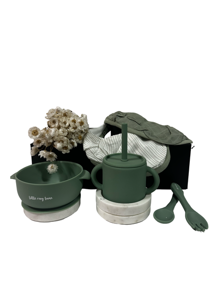 A set of baby mealtime essentials including a green silicone bib, bowl, cup, and cutlery, presented with a white magnetic gift box and decorative white flowers.
