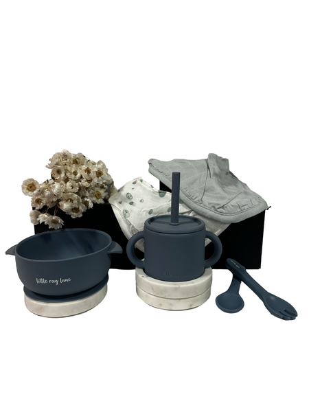 A set of baby mealtime essentials including a green silicone bib, bowl, cup, and cutlery, presented with a white magnetic gift box and decorative white flowers.