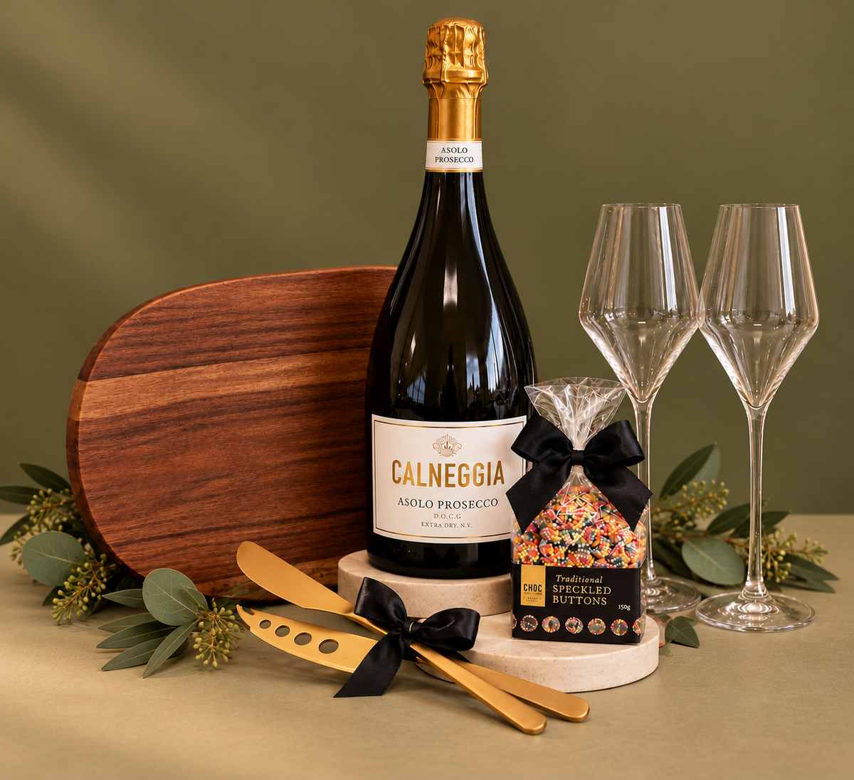 A luxury champagne gift set featuring a bottle of Caleggia Asolo Prosecco, two clear champagne flutes, a wooden cutting board, gold-toned cheese knives, and a bag of gourmet cheese sticks, all arranged with black gift boxes, a black ribbon, and holiday greenery on a white background.
