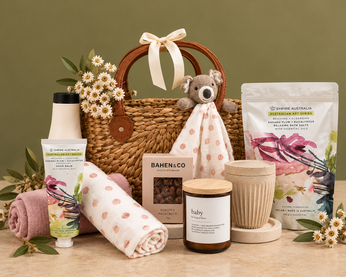 A curated self-care and baby gift basket arrangement on a wooden surface, featuring a woven basket with a bunny plush, a tube of hand lotion, a rolled towel, a candle, a mug, bath salts, and a baby blanket, styled with green foliage and eucalyptus.