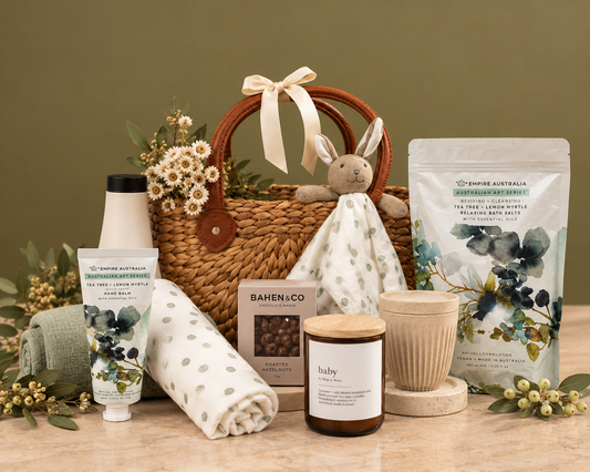 A curated self-care and baby gift basket arrangement on a wooden surface, featuring a woven basket with a bunny plush, a tube of hand lotion, a rolled towel, a candle, a mug, bath salts, and a baby blanket, styled with green foliage and eucalyptus.