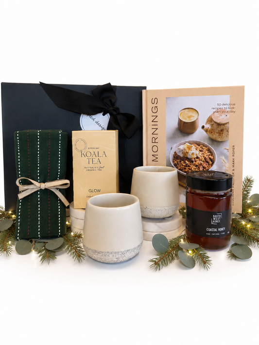 A curated gift set arranged on a white surface with a black gift box, a dark green striped tea bag bundle, a box of Koala Tea, a cookbook titled Mornings, two cream ceramic mugs, and a candle jar with a black label.