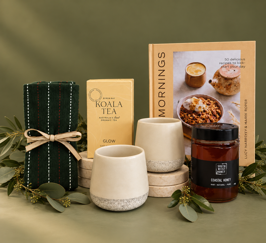 A curated gift set arranged on a white surface with a black gift box, a dark green striped tea bag bundle, a box of Koala Tea, a cookbook titled Mornings, two cream ceramic mugs, and a candle jar with a black label.