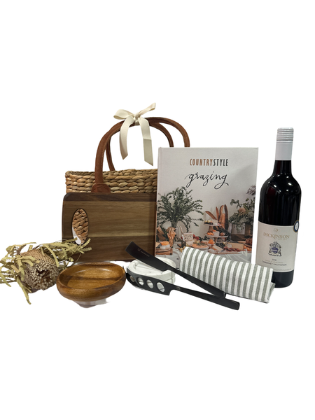 A gift basket containing a wooden paddle board, a wooden bowl, a set of black cheese knives, a bottle of Dickinson Estate Shiraz, and a green and white striped tea towel, all arranged on a white background.