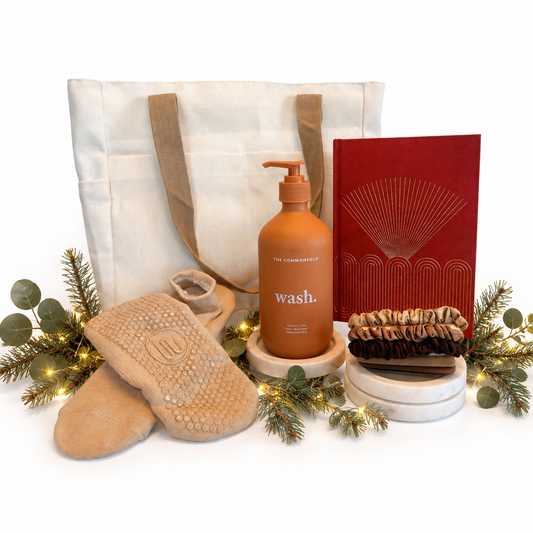 A curated gift set featuring a cream canvas tote bag, a terracotta pump bottle labeled 'wash.' by The Commonfolk Collective a red notebook with gold line art, a stack of brown scrunchies, and tan textured footie socks, styled with eucalyptus branches and fairy lights on a white background.