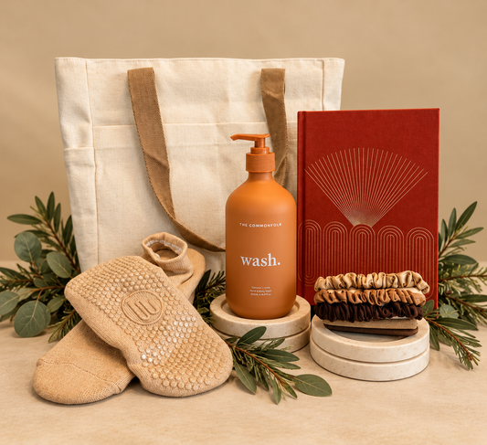 A curated gift set featuring a cream canvas tote bag, a terracotta pump bottle labeled 'wash.' by The Commonfolk Collective a red notebook with gold line art, a stack of brown scrunchies, and tan textured footie socks, styled with eucalyptus branches and fairy lights on a white background.