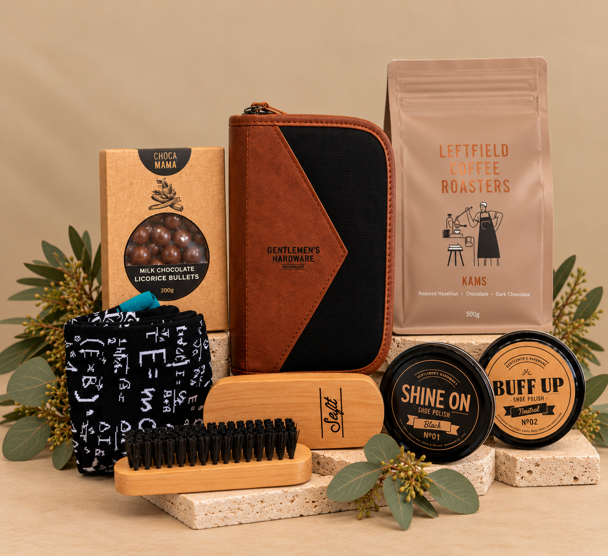 A curated gift set arranged on a white background with pine branches, eucalyptus, and fairy lights. The set includes a black gift box with a black ribbon, a brown leather and black zip pouch, a bag of Leftfield Coffee Roasters, a black wallet, a small round tin labeled Shine On, a black item labeled Buff Up, a black scrubber, a wooden brush, and a small packaged accessory.