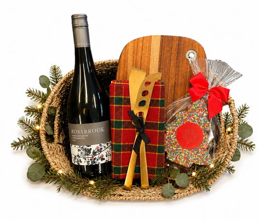 A holiday gift basket arrangement on a white background, featuring a Rosabrook wine bottle, a wooden cutting board, a red plaid cheese holder, wooden serving utensils, and a decorated cookie with a red bow.