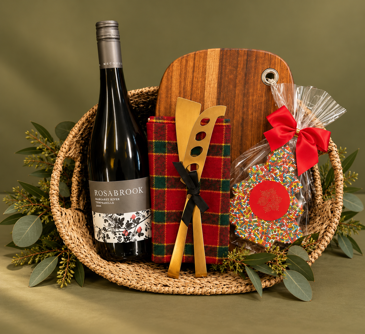 A holiday gift basket arrangement on a white background, featuring a Rosabrook wine bottle, a wooden cutting board, a red plaid cheese holder, wooden serving utensils, and a decorated cookie with a red bow.