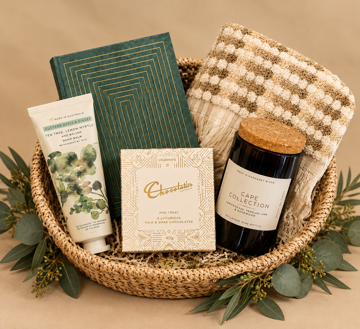 A curated gift basket arrangement in a woven round basket, featuring a green geometric notebook, a cream bath and body lotion tube, a chocolate-covered treat box, a dark candle jar with a cork lid, and a cream-and-brown checkered throw blanket. The arrangement is decorated with pine branches, eucalyptus leaves, and warm string lights on a white background.