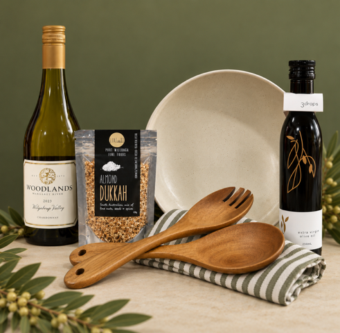 Bottle of wine, cream salad bowl, bottle of oil, packet of dukkah, wooden salad servers and striped tea towel