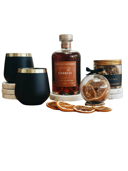Starward Whisky bottle with black stemless glasses, dried oranges, and a jar of nuts on a white background