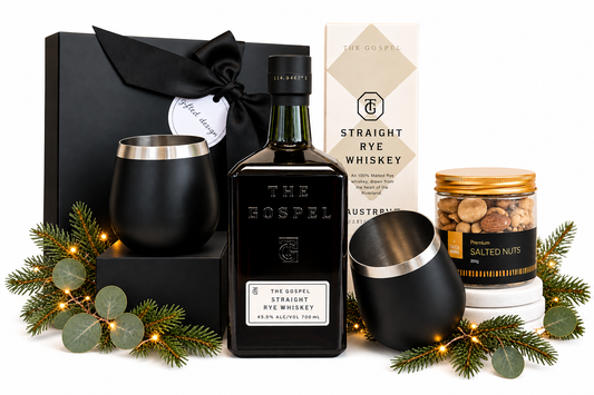 A curated gift set featuring a bottle of The Gospel Straight Rye Whiskey, a black tumbler, a black gift box with a ribbon, a candle, a jar of salted nuts, and a box of The Gospel Straight Rye Whiskey, styled with eucalyptus and fairy lights.