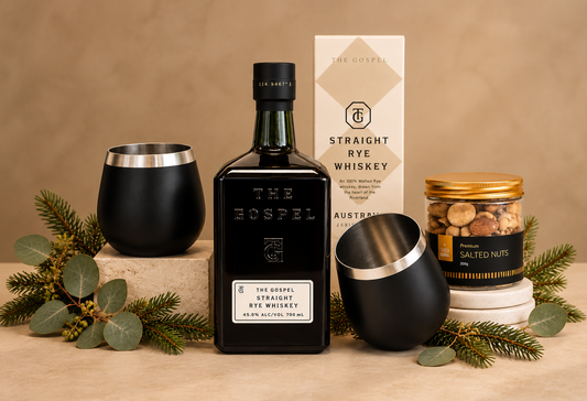 A curated gift set featuring a bottle of The Gospel Straight Rye Whiskey, a black tumbler, a black gift box with a ribbon, a candle, a jar of salted nuts, and a box of The Gospel Straight Rye Whiskey, styled with eucalyptus and fairy lights.