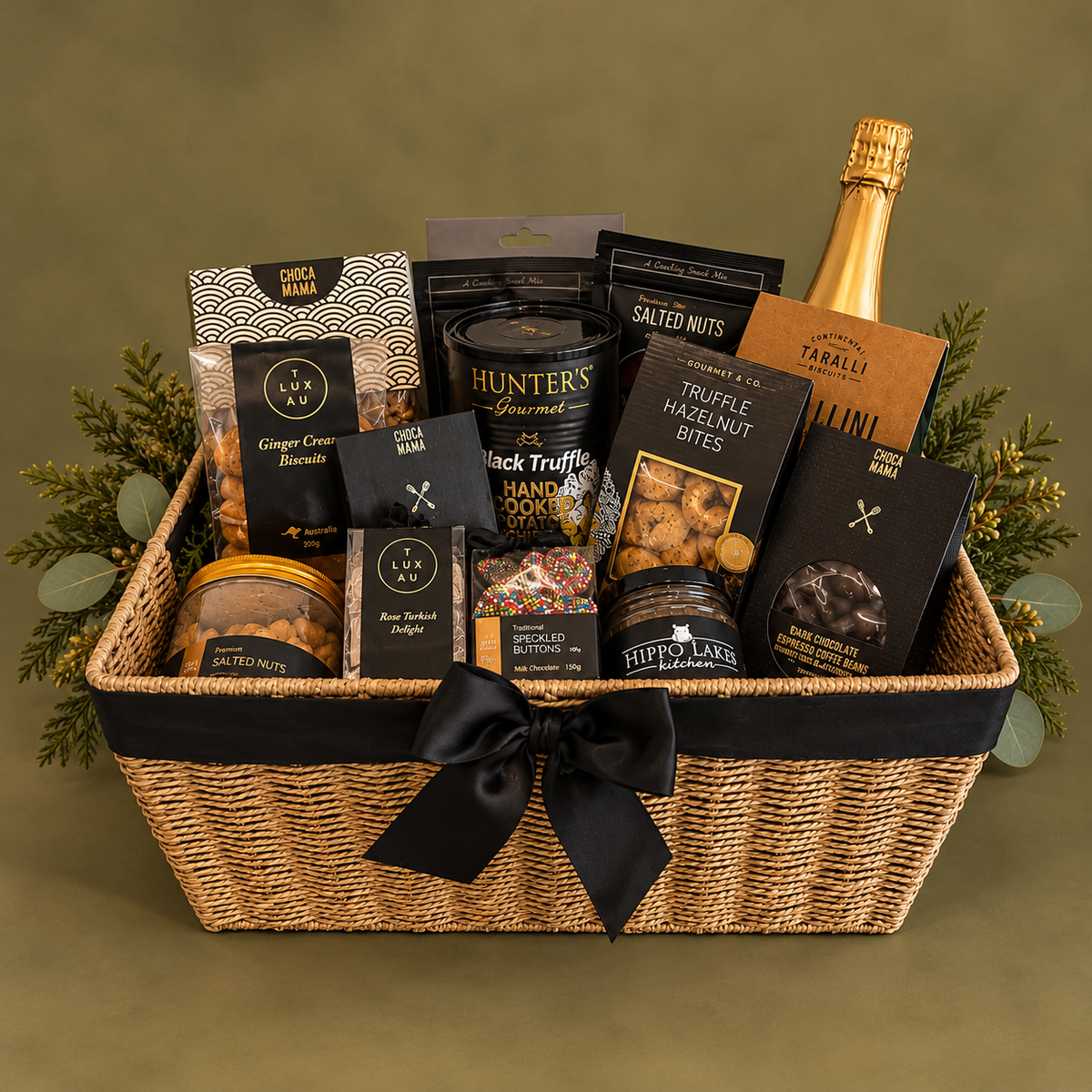 A gourmet gift basket filled with assorted snacks, nuts, and a bottle of champagne, arranged in a woven straw basket with a black ribbon and bow. Visible brands include Hunter's Gourmet, Truffle Farms, and Farrow & Ball.