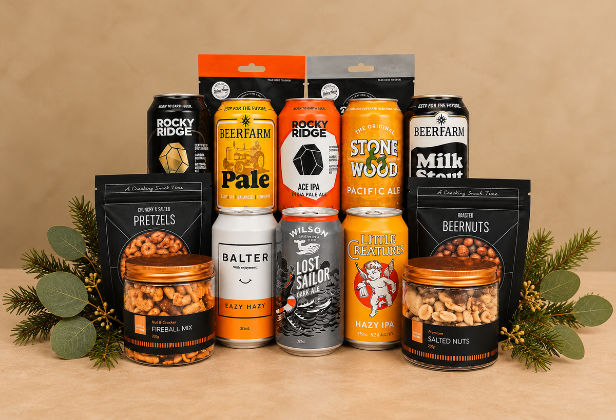 A holiday gift bundle of craft beer, pretzels, and nuts arranged on a white background with pine branches and fairy lights. The bundle includes cans and bags from Rocky Ridge, Beerfarm, Stone Wood, Little Meats, Wilson's, and Balter.