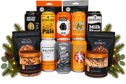 A holiday gift bundle of craft beer, pretzels, and nuts arranged on a white background with pine branches and fairy lights. The bundle includes cans and bags from Rocky Ridge, Beerfarm, Stone Wood, Little Meats, Wilson's, and Balter.