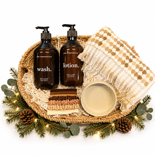 A curated spa gift set arranged in a woven round tray on a white background, surrounded by pine branches, eucalyptus, pinecones, and fairy lights. The set includes two dark amber pump bottles labeled 'wash.